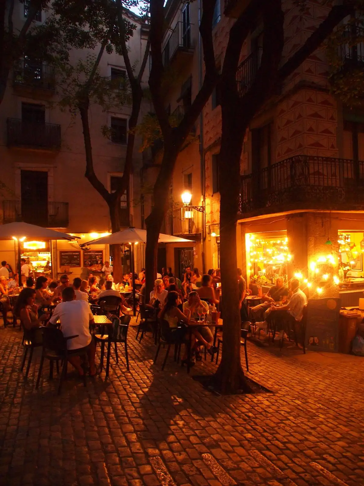 Terrassa plena de gent en una plaça de casc antic al vespre, amb els llums càlids d'un bar al fons.