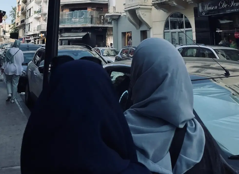 Dues persones amb hijab vistes per darrere en un carrer ple de cotxes i botigues.