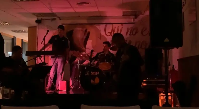 Un grup toca en un escenari petit amb llum vermellosa: teclats, bateria i baix davant un mural pintat.