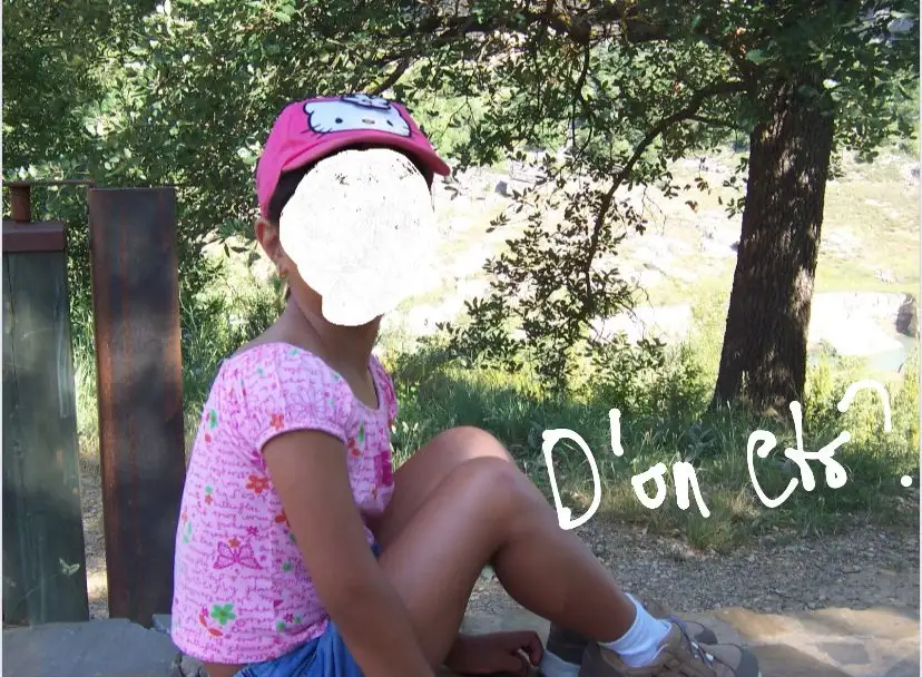 Una nena asseguda en una roca del bosc amb gorra rosa i la cara difuminada; al costat, escrit a mà, «D'on ets?».