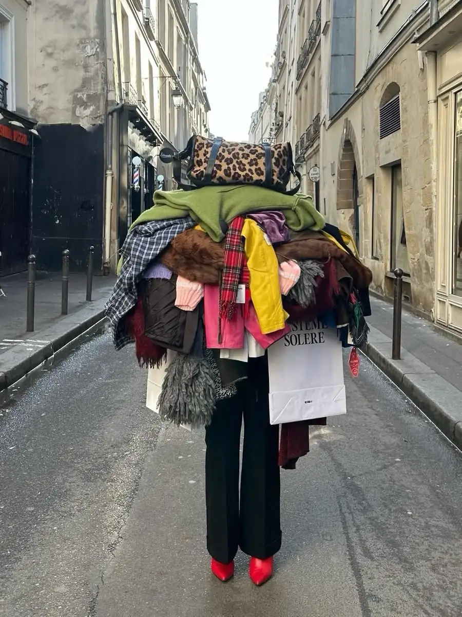 Una persona travessa un carrer carregada amb una muntanya de roba i bosses que li tapa tota la cara.