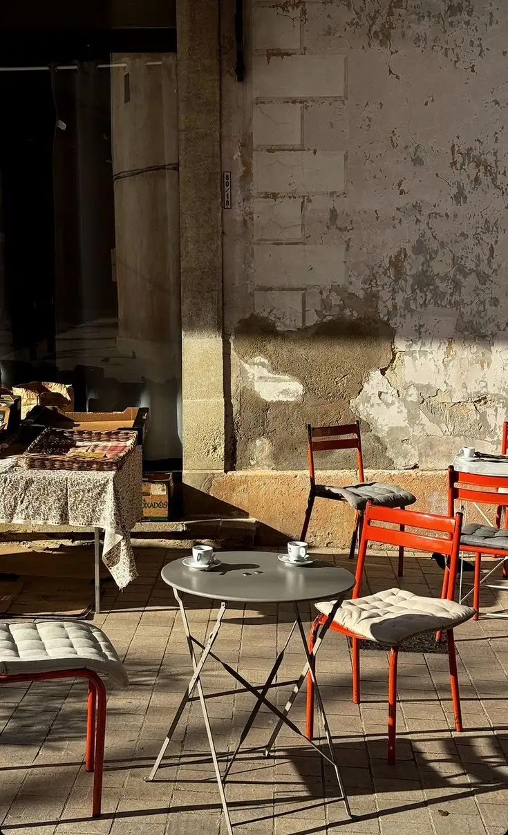 Terrassa buida d'un cafè amb cadires taronges i dues tasses sobre una taula rodona, il·luminada pel sol baix.