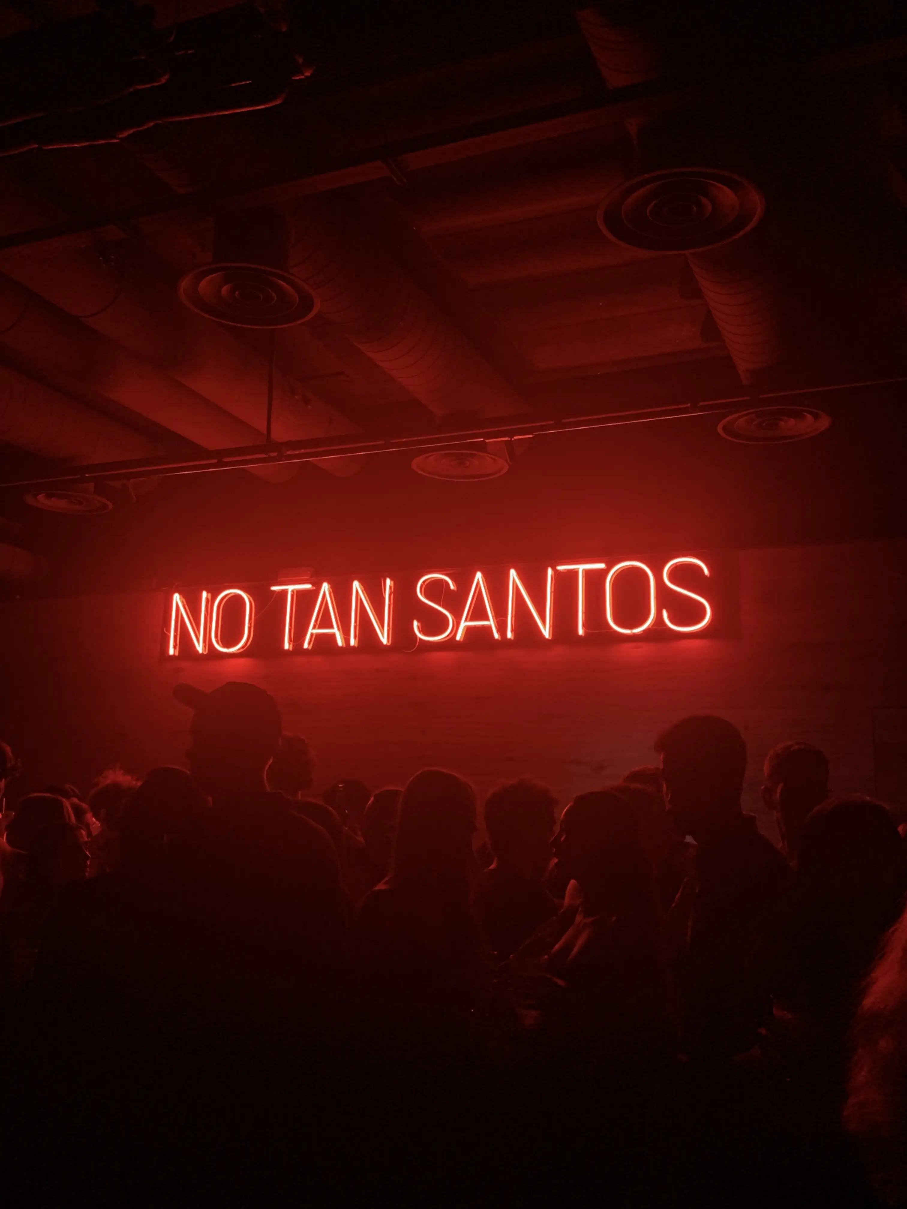 Rètol de neó vermell amb el text «No tan santos» il·luminant una sala fosca plena de gent.